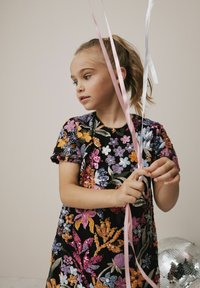 Schwarzes Kleid mit lebhaften, gestickten Blumenmustern und Pailletten. Kind hält rosa und weiße Bänder. Eine Discokugel ist in der Nähe.