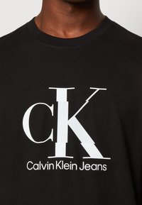 Černé tričko s velkým bílým grafickým prvkem s textem "CK" a "Calvin Klein Jeans". Vyrobeno z bavlny, s kulatým výstřihem a krátkými rukávy.