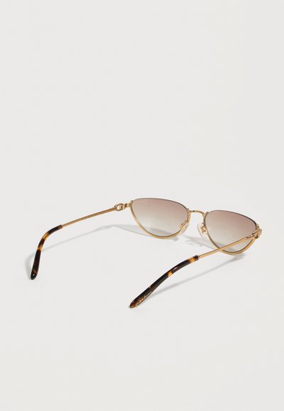 Γυαλιά ηλίου με χρυσό σκελετό σε σχήμα γατιών, με βαθμιαίους καφέ φακούς και άκρα βραχίονα με σχέδιο χελώνας, που ακουμπούν σε λευκή επιφάνεια.