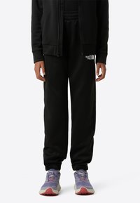 Čierne tepláky s elastickým pásom a manžetami, s malým bielym logom. Nosené s fialovými a smotanovými športovými teniskami.