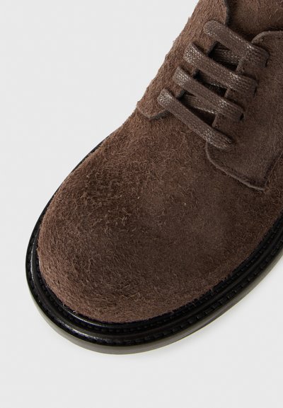 Zapato de ante marrón con un acabado texturizado, puntera redonda y suela de goma negra. Presenta detalles de cordones y costura acabada alrededor del borde.