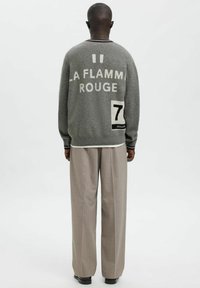 Pelēks vilnas džemperis ar uzrakstu "LA FLAMME ROUGE" baltā krāsā un melnu un baltu 7. numura uzplākšņu. Kombinēts ar gaiši bēšām biksēm.