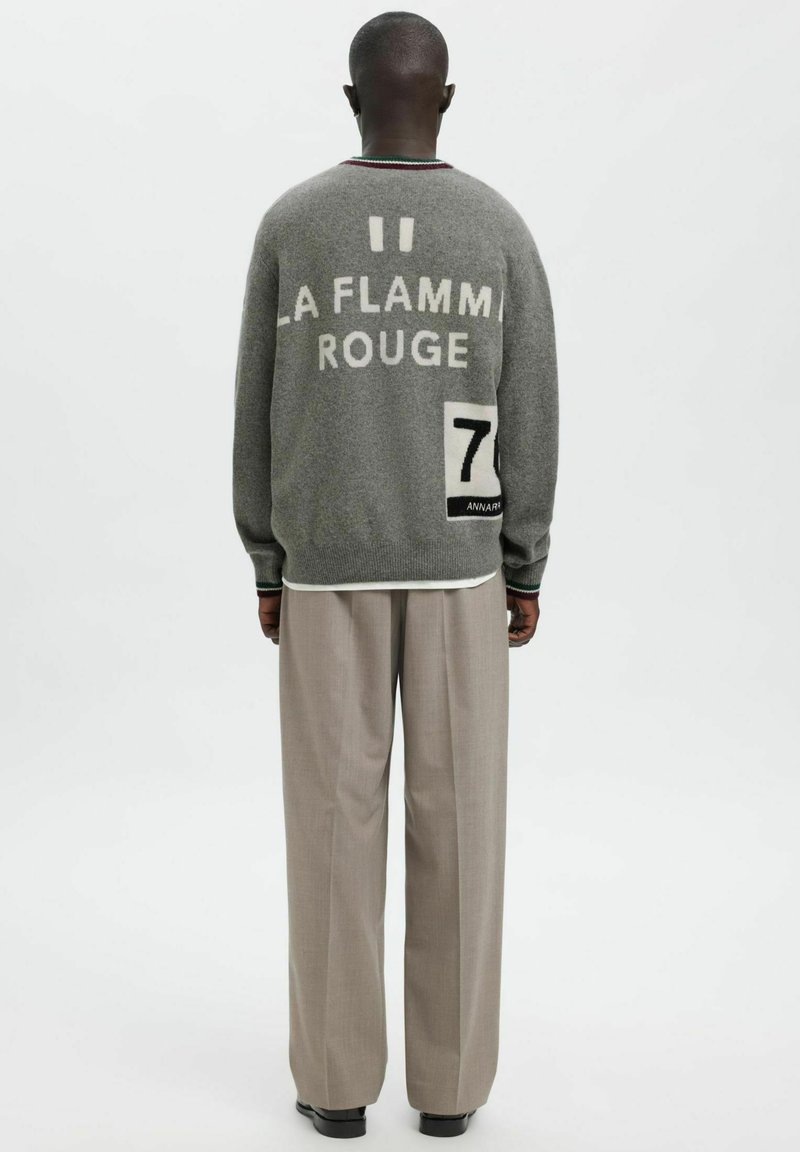 Pelēks vilnas džemperis ar uzrakstu "LA FLAMME ROUGE" baltā krāsā un melnu un baltu 7. numura uzplākšņu. Kombinēts ar gaiši bēšām biksēm.
