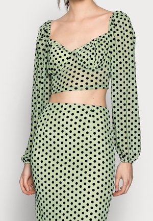 Kvinde iført en lysegrøn cropped top og en højtaljet nederdel, begge med sorte polkaprikker; toppen har lange puffede ærmer og hjerteformet udskæring.