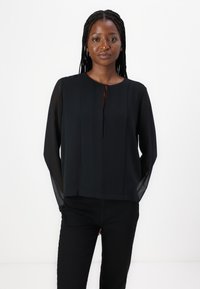 Top leggero nero con maniche lunghe e trasparenti, scollo rotondo, pieghe sottili e dettaglio a chiave sul davanti. Indossato con pantaloni neri.