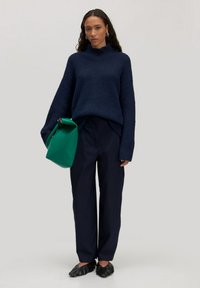 Marineblaue Strickjacke mit hohem Kragen, schwarze weit geschnittene Hose, grüne Tasche mit strukturiertem Design und schwarze flache Schuhe mit Rüschen.