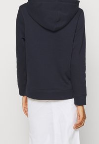 Personne portant un sweat à capuche bleu marine et une jupe blanche, vue de dos avec les bras relâchés sur les côtés.