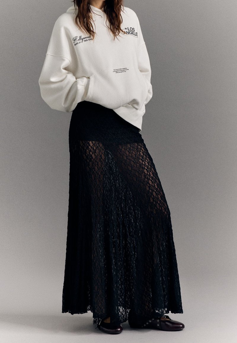 Felpa bianca oversize con stampa, abbinata a una gonna maxi nera in pizzo con un design a strati trasparente. Scarpe nere con dettagli hardware.