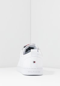 Baskets en cuir blanc avec une forme arrondie, semelle plate, talon avec tirant, et un petit logo en rouge, blanc et bleu à l'arrière.