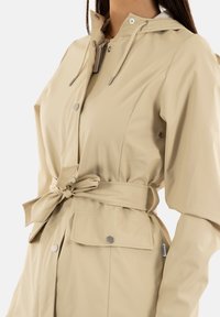 Veste imperméable beige avec taille froncée, capuche et boutons-pression. Présente une texture lisse, des poches latérales et un détail de ceinture à nouer.