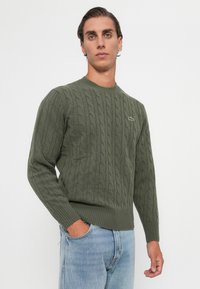 Mann iført olivengrønn kabelstrikket genser med Lacoste-logo og lyseblå denimjeans, stående med den ene hånden i lommen.