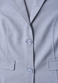 Blazer gris clair avec deux boutons visibles et poches à rabat droites, présentant un tissu finement texturé et des coutures ajustées.