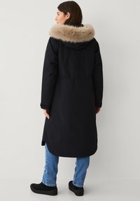 Parka nera con cappuccio in pelliccia sintetica, maniche lunghe e taglio al ginocchio. Presenta una vita aderente e una texture liscia. Indossata con jeans blu.