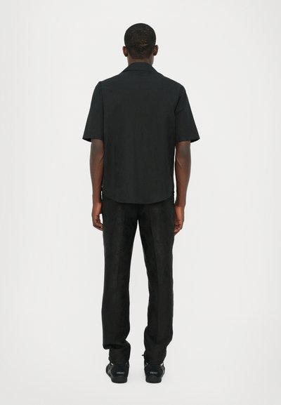 Chemise noire à manches courtes avec un ourlet droit, associée à un pantalon noir ajusté avec une texture à motif subtil, portée avec des baskets noires.