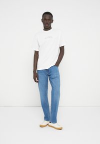 Νεαρός άνδρας στέκεται φορώντας λευκό μπλουζάκι με κείμενο, μπλε τζιν και λευκά αθλητικά παπούτσια με καφέ σόλες, πάνω σε απλό λευκό φόντο.