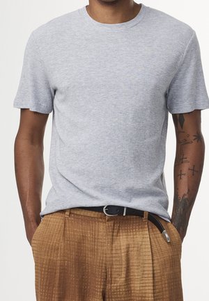 Mężczyzna w jasnoszarej, krótko rękawowej dzianinowej koszuli włożonej do wysokich, brązowych, teksturowanych spodni z czarnym paskiem, z widocznymi tatuażami na ramionach.