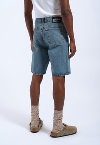 Shorts in denim di colore azzurro, con taglio dritto e design a cinque tasche. Indossati con sandali beige e calzini lavorati a maglia. Etichetta visibile in vita.