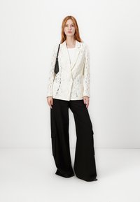 Lace blazer in cream with a tailored design, featuring a single-button closure and two front pockets, paired with wide black trousers.