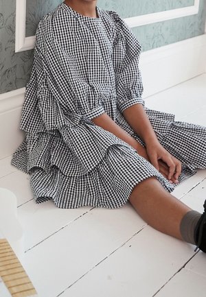 Robe noire et blanche à motif vichy avec volants en étages, manches bouffantes et encolure ronde, posée sur un sol en bois clair.