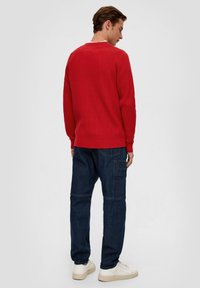 Rode geribde trui met lange mouwen, gedragen over vervaagde blauwe spijkerbroek. De outfit wordt compleet gemaakt met lichtgekleurde sneakers.