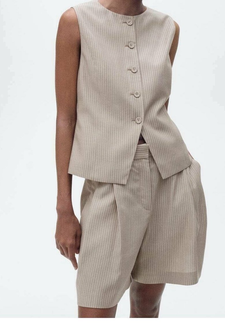 Gilet beige a righe con bottoni rotondi e pantaloni corti coordinati. Entrambi hanno una vestibilità sartoriale e una texture sottile, creando un outfit coeso.