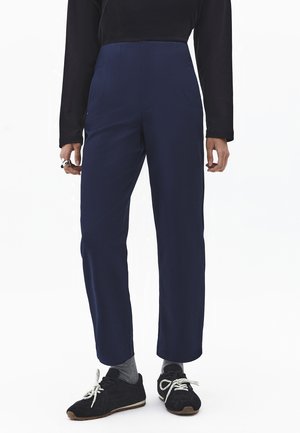 Persoon draagt een marineblauwe broek met hoge taille, een zwart shirt met lange mouwen, grijze sokken en zwarte sneakers met witte veters, staand tegen een witte achtergrond.