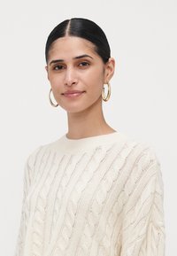 Woman with dark hair in a low bun wearing gold hoop earrings and a cream cable-knit sweater, against a plain white background.