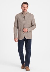 Braune Tweedjacke mit Stehkragen, zwei Vorderaschen und Knopfschluss. Kombiniert mit Jeanshosen und braunen Schuhen.