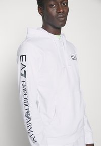 Felpa bianca con cappuccio, dotata di una tasca frontale e maniche lunghe. "EA7 Emporio Armani" stampato in nero lungo la manica sinistra e sul petto.