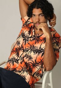 Korte mouwen shirt met een tropisch patroon; oranje en crèmekleurige achtergrond met zwarte silhouetten van palmboompjes; lichtgewicht stof, kraagdetail.