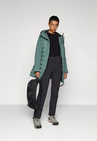 Jakku jadranske plave boje s postavom, crna majica i uske crne hlače. Nosite sive planinarske čizme i držite crni ruksak. Jednostavan dizajn, funkcionalan izgled.