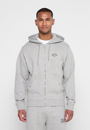 Mann trägt hellgrauen Kapuzenpullover mit Reißverschluss und passende Jogginghose, steht vor einfachem weißen Hintergrund.