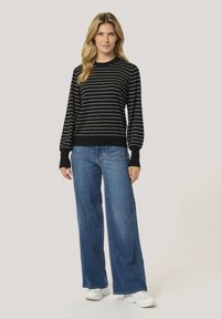 Sweter w czarno-białe paski z długimi rękawami, z prążkowanymi mankietami, zestawiony z szerokimi niebieskimi dżinsami i białymi sneakersami. Gładkie tło.
