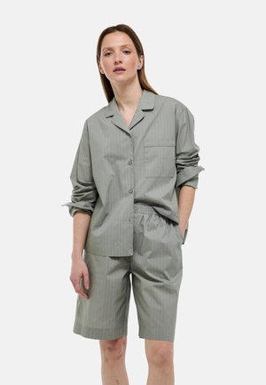 Femme portant une chemise à rayures fines gris clair, ample, boutonnée, avec les manches retroussées, et un short assorti arrivant au genou, sur fond blanc.