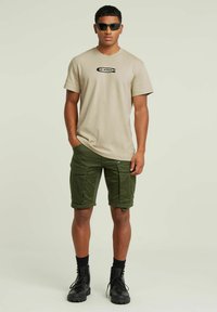 T-shirt beige in cotone con logo nero, abbinata a pantaloni cargo verdi con tasche laterali e taglio al ginocchio. Stivali da combattimento neri.