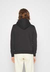 Felpa con cappuccio nera in un tessuto testurizzato, con polsini e orlo a coste. La vista posteriore mostra una tasca a marsupio e un design semplice.