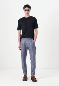 T-shirt noir à manches courtes, pantalon plissé gris clair et mocassins marron. Modèle portant des lunettes de soleil noires, fond neutre.