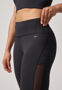 Leggings noirs taille haute avec des panneaux en maille sur les côtés, fabriqués à partir d'un matériau extensible. Présente un logo sur la ceinture et des détails de couture subtils.