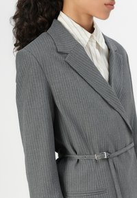 Grå striped blazer med en skreddersydd design, som har krage, belte i midjen og en enkelt lomme foran.