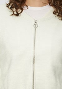 Veste zippée crème clair avec tissu texturé, encolure ronde et fermeture éclair avec détails en métal argenté.