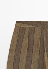 Brune og beige stribede linnedbukser med højtaljet pasform og lodrette folder, synlige på en hvid baggrund.