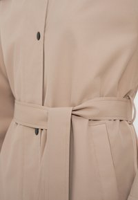 Manteau beige ceinturé en tissu lisse, manches longues, boutons à l'avant et poches latérales. Accentué par des boutons noirs et une ceinture à nouer.
