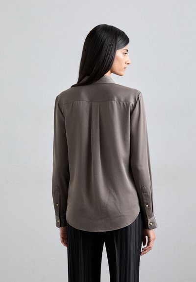 Long-sleeve shirt in dark taupe fabric, features a shirt collar, smooth texture, back yoke detail, and buttoned cuffs. Paired with black pleated trousers.