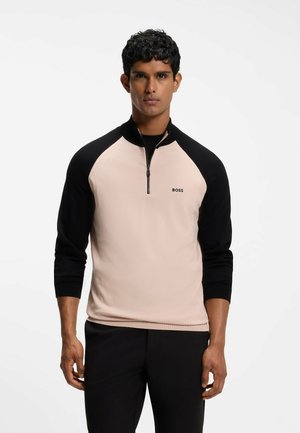 Homme debout face à l'objectif portant un pull à demi-zip beige et noir à manches longues avec le logo "BOSS", pantalon noir, fond neutre.
