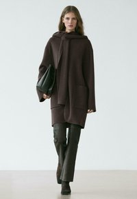 Manteau en laine marron avec une grande capuche, des poches frontales et une texture lisse, associé à un pantalon en cuir ajusté et un sac noir.