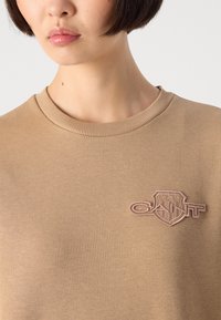 Giovane con i capelli corti e scuri che indossa un maglione a girocollo beige con un logo GANT ricamato in rosa sul petto.