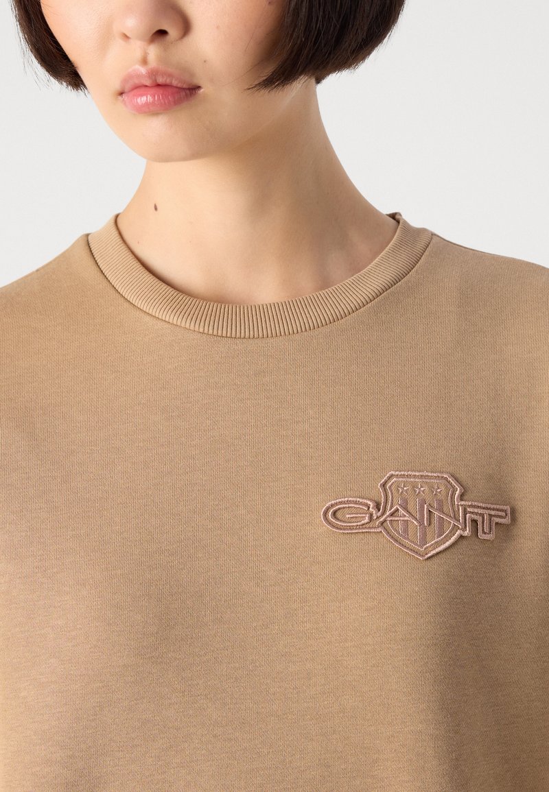 Junge Person mit kurzem dunklem Haar, die einen beigen Rundhals-Pullover mit einem rosa gestickten GANT-Logo auf der Brust trägt.