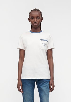 Kvinde med fletninger, iført en hvid t-shirt med korte ærmer, blå kant omkring kraven og lynlås på brystlommen, kombineret med blå jeans.