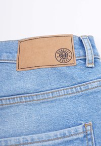 Ljusblå denimjeans med en texturerad läderpatch som visar "ÄRLIGHET ÄR REGELN." Har bruna sömmar och en bakficka.