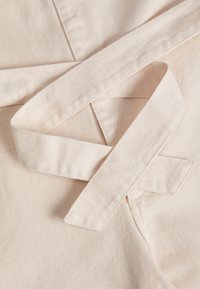 Tejido de algodón beige con una textura acanalada, que presenta un lazo anudado. El tejido es suave, con costuras visibles y un acabado limpio.